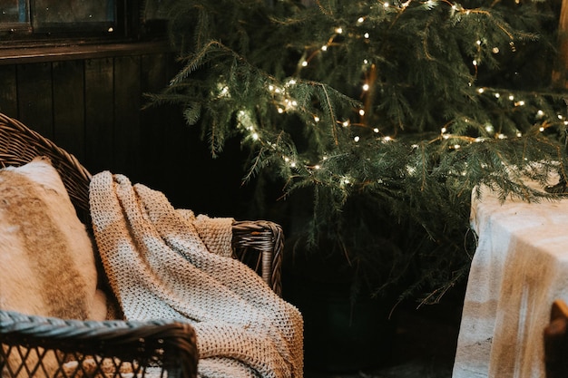 Christmas and new year time cozy armchair or wicker easy chair with a comfy knitted plaid and a fur pad pillow near a decorated christmas tree with glowing light winter holiday season at home