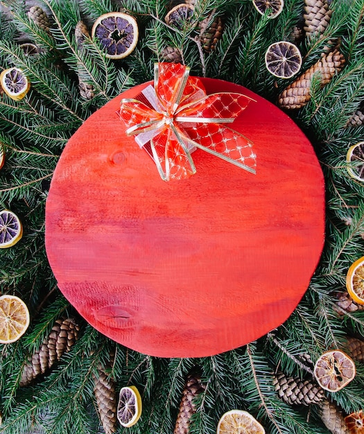Рождество, новогодний зимний праздничный состав с красной деревянной поверхностью на зеленых еловых ветвях, высушенными апельсиновыми дольками, шишками и красным бантом.