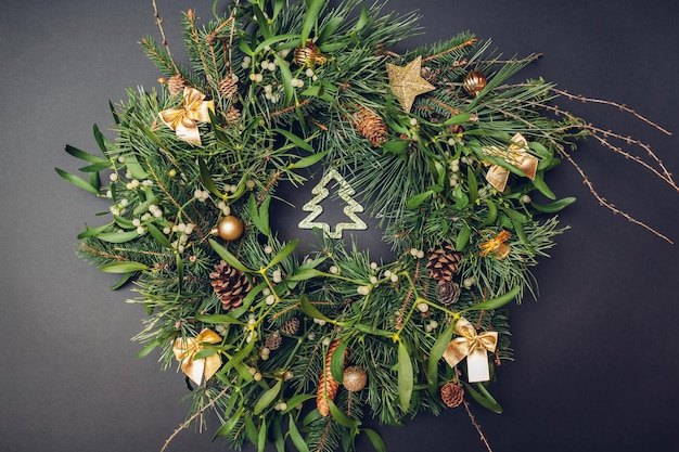 Christmas and New Year wreath made of fir, pine, mistletoe branches, cones decorated with golden ornaments, toys, bows on grey background.