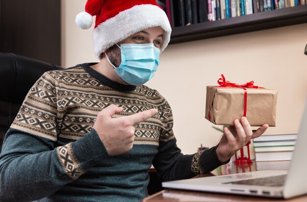 Christmas online congratulation. Young man in santa claus hat and face mask gives a gift and talks using laptop for video call friends and childrens. Christmas during coronavirus.