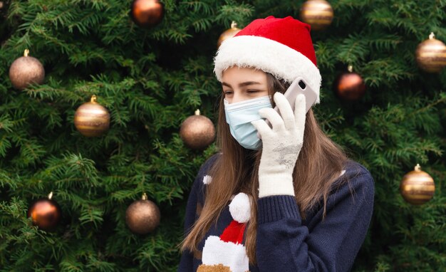 Christmas online greetings. Close up Portrait of woman wearing a santa claus hat and medical mask with emotion. Against the background of a Christmas tree. Coronavirus pandemic