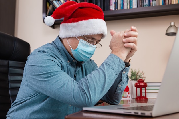 Christmas online prayer. Senior man in santa claus hat prayer using laptop for video call friends and childrens. The room is festively decorated. Christmas during coronavirus.