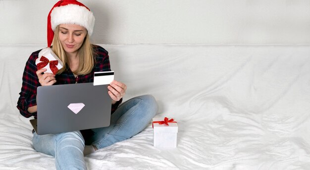 Christmas online shopping. Female buyer making order on laptop holding card and gift box.