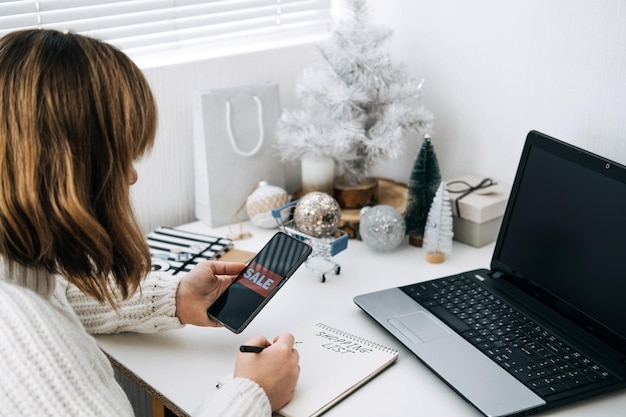 クリスマスオンラインショッピング冬のセールブラックフライデーの女性とショッピングリストのスマートフォンとラップトップ