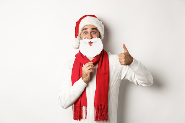 Рождественская вечеринка и концепция празднования. Счастливый мужской модели в шляпе Санта-Клауса и белой маске бороды, показывая большой палец вверх жест, стоя на белом фоне