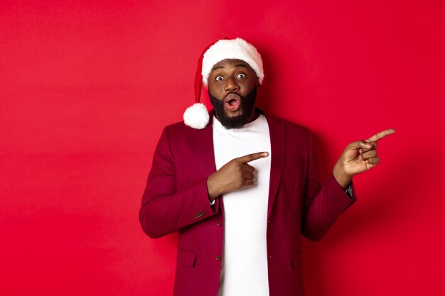 クリスマス、パーティー、休日のコンセプト。興奮した黒人男性が広告を表示し、プロモーションオファーで指を指して、赤い背景にサンタの帽子をかぶって立っている