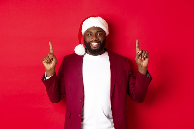 Christmas, party and holidays concept. Handsome african american man in santa hat smiling, pointing fingers up and showing advertisement, standing over red background