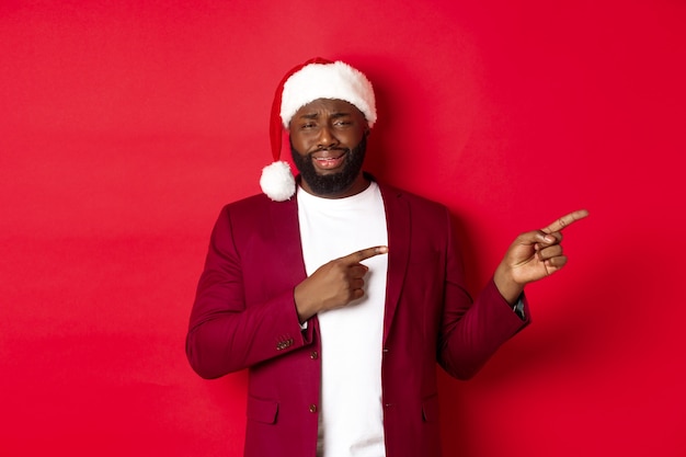 Christmas, party and holidays concept. Skeptical and unamused Black man looking with disdain, pointing fingers right at logo, standing in santa hat against red background