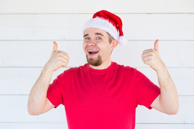 Рождество и люди концепции - веселый молодой человек в рождественской шляпе показывает палец вверх на белом пространстве.