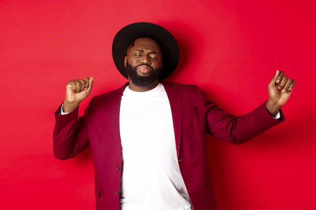 Christmas and people concept. Happy Black man celebrating New Year and dancing, wearing party outfit, red background.