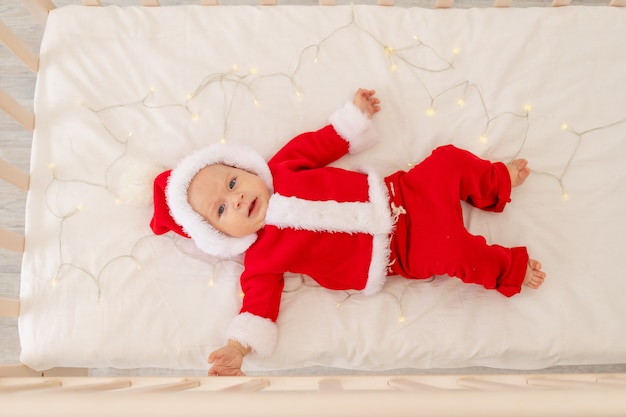 自宅のベビーベッドで横になっているサンタスーツの赤ちゃんのクリスマス写真