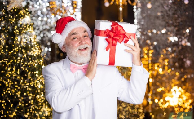 Christmas preparation. senior man santa hat hold gift box. bearded businessman present. holiday decor illumination. stay happy this christmas. happy new year. cheap xmas present and business reward.