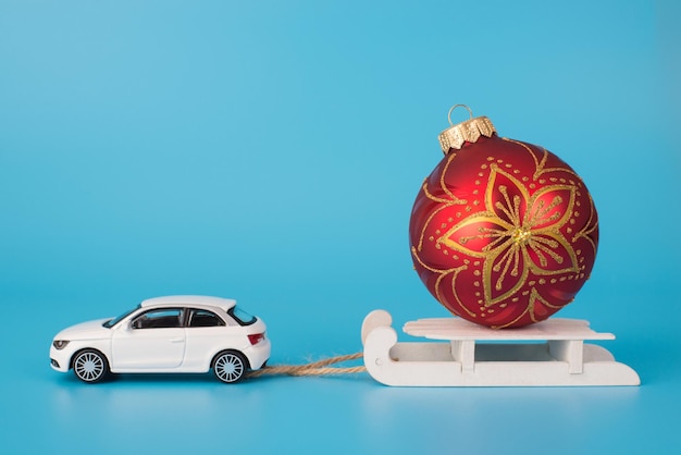 Christmas preparations concept. Close up photo of toy mini car carrying white wooden sledge with red golden ornament bauble on top isolated on blue background