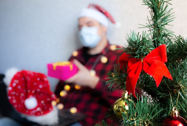 Christmas presents giving. Man in medical mask sharing box gifts through video calls. Holidays in isolation. New year tree and lights with decorations. Family and friends celebrating online.