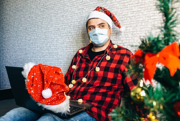 Regali di natale che danno. uomo in maschera medica che condivide regali tramite videochiamate. vacanze in isolamento. albero di capodanno e luci con decorazioni. famiglia e amici che festeggiano online.