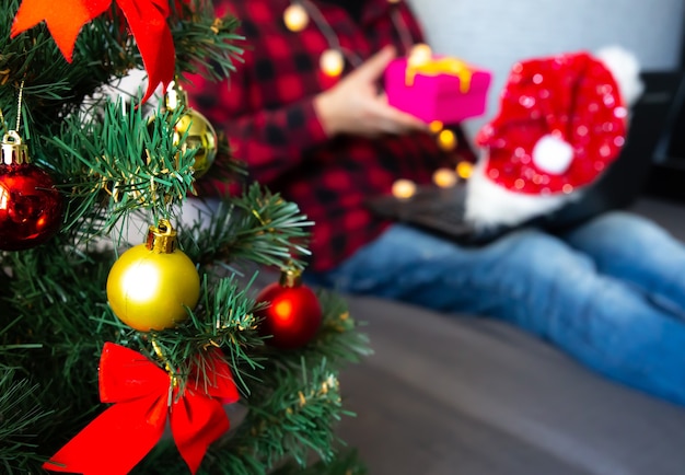 Christmas presents giving. Man sharing box gifts through video calls. Holidays in isolation. New year tree and lights with decorations. Family and friends celebrating online.