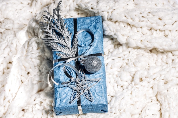 Christmas presents with gift box in silver blue color decorated with pine cones and twigs on white knitted plaid, preparation for holidays. Christmas presents and New Year. Handmade. Selective focus.
