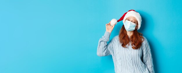 写真 クリスマスの検疫とコビッドのコンセプト サンタの帽子と顔のマスクを見つめる陽気な10代の赤毛の女の子