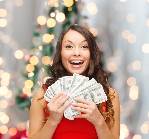 Concetto di natale, vendita, bancario, vincente e vacanze - donna sorridente in abito rosso con soldi in dollari americani su sfondo soggiorno e albero di natale