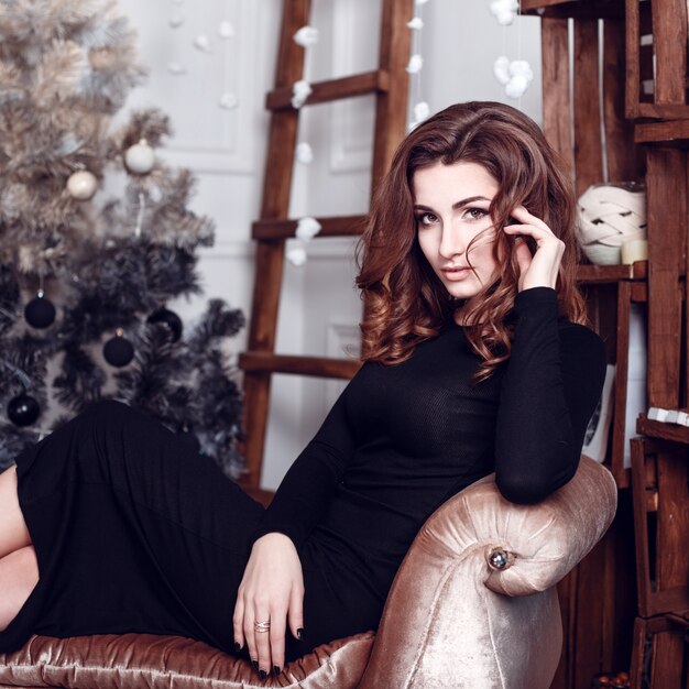 Christmas Santa. Beautiful smiling woman model. Makeup. Healthy long hair style. Elegant lady in black dress over christmas tree lights background. happy new year