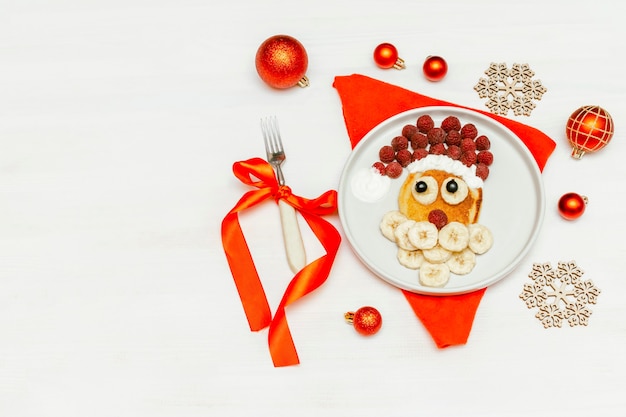 Christmas Santa Claus-gezichtsvormige pannenkoek met zoete verse frambozenbes en banaan op plaat op witte achtergrond voor het ontbijt van jonge geitjeskinderen. xmas food dessert, nieuwjaarsdecoratie, vork, servet.