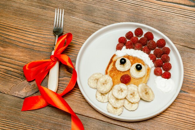 Christmas santa claus vormige pannenkoek met zoete verse frambozenbes en banaan op plaat op houten achtergrond voor kinderen kinderen ontbijt. xmas eten met nieuwjaarsdecoraties close-up