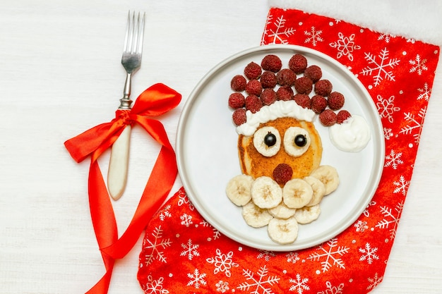 Christmas Santa Claus vormige pannenkoek met zoete verse frambozenbes en banaan op plaat, rode sok, vork op witte houten achtergrond voor kinderen kinderen ontbijt. kerstmaaltijd met nieuwjaarsversieringen