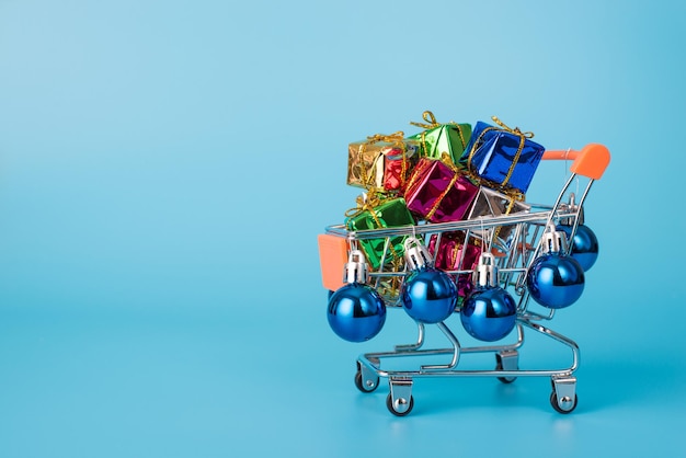 Christmas shopping concept. close up photo of toy shopping cart with pile of giftboxes with little blue baubles isolated on blue background