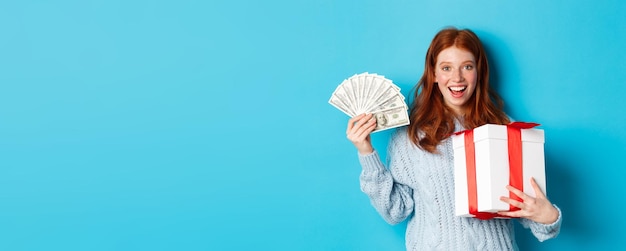 Photo christmas and shopping concept happy redhead woman holding money and big xmas present showing dollar