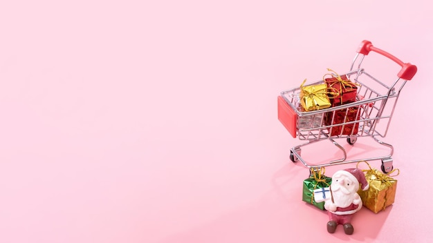 Christmas shopping concept mini red shop cart trolley with Santa Claus toy and gift box isolated on pale pink background blank copy space close up