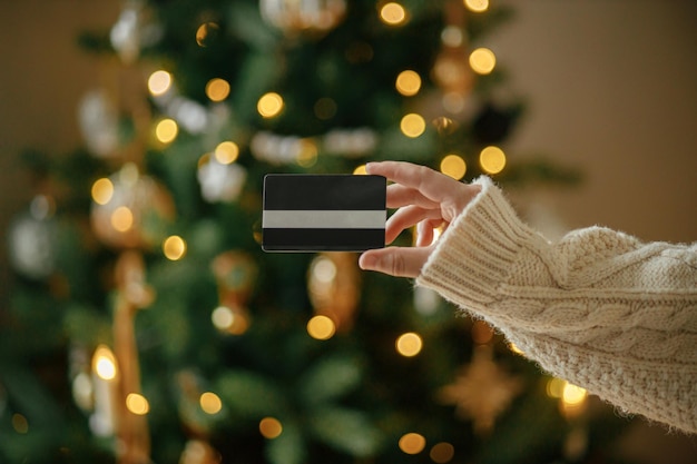 Christmas shopping online and black friday sales Hand in cozy sweater holding credit card close up on background of christmas tree lights in festive decorated room