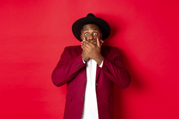 Christmas shopping and people concept. shocked black man looking startled at camera, covering mouth with hands, standing against red background