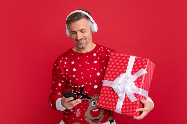 Christmas songs santa with phone headphone and gift studio\
portrait of guy wears santa hat and winter sweater man with\
christmas hat over isolated red background