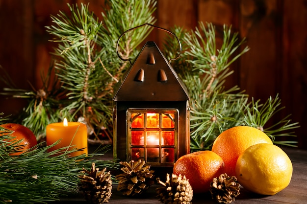 Рождественский натюрморт с фонарем, горящая свеча, ветви елки, фрукты и сосновые шишки.