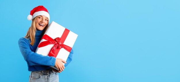 Photo christmas surprise happy lady in santa hat embracing big gift box on blue studio background panorama