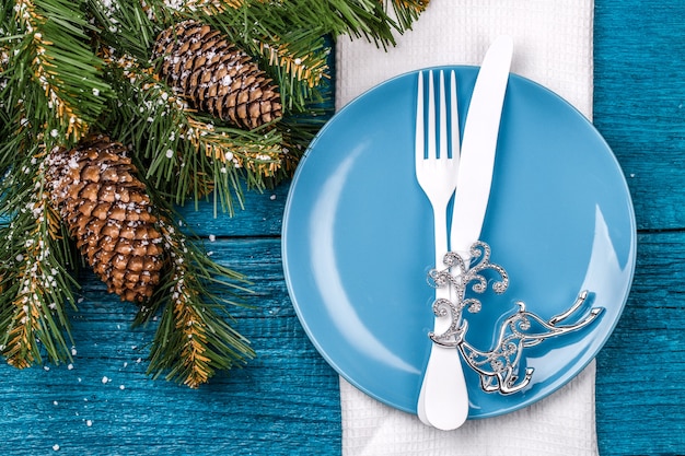 크리스마스 테이블 장소 설정-흰색 냅킨, 파란색 접시, 흰색 포크와 나이프, 장식 된 크리스마스 트리 장난감 파란색 테이블-실버 사슴과 크리스마스 소나무 가지. 크리스마스 휴일 배경입니다.
