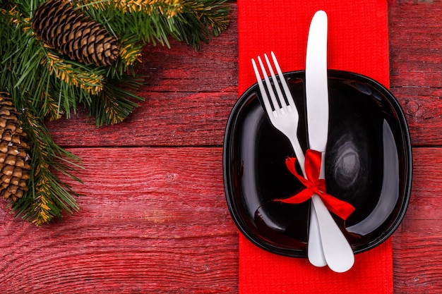 Сервировка рождественского стола с красной салфеткой, черной тарелкой, белой вилкой и ножом, украшенным красным бантом и рождественскими сосновыми ветками. Стол рождественских праздников.