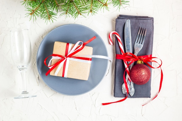 Christmas table place setting with tree fir branches, napkin, fork, knife, ball, plate, candy canes, gift and glass.