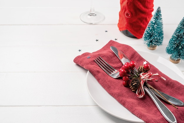 Christmas table setting with white dishware silverware and red and green decorations on white wooden background