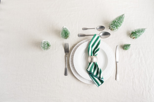 Christmas table setting with white plates, cutlery, napkin and with Christmas decor on linen tablecloth with copy space. Winter, festive concept table serving for New year.