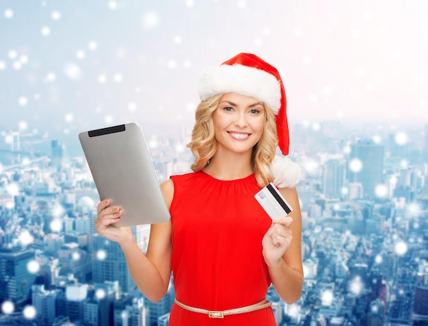 Christmas, technology, shopping and people concept - smiling woman in santa helper hat with tablet pc computer and credit card over snowy city background