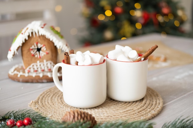 christmas time, two white cup of cocoa with marshmallows and gingerbread house on table