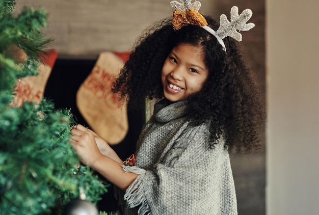 Christmas tree and decorating with a girl in antlers putting an ornament on a branch in the living room of her home Children portrait and smile with a happy kid placing decorations for celebration