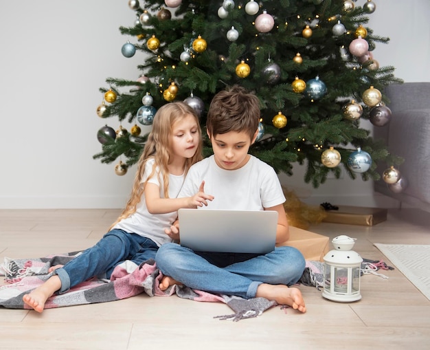 Albero di natale in un ampio soggiorno. i bambini giocano vicino all'albero di natale. un ragazzo con un laptop comunica con gli amici.