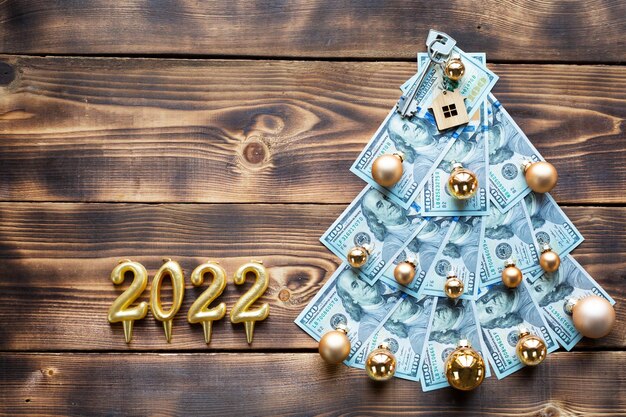 Foto albero di natale fatto di banconote da 100 dollari e chiave di casa, candele con i numeri 2022. decorazioni natalizie di finanza, risparmio, ricchezza, spese nel nuovo anno. immobili, abitazioni, mutui, traslochi