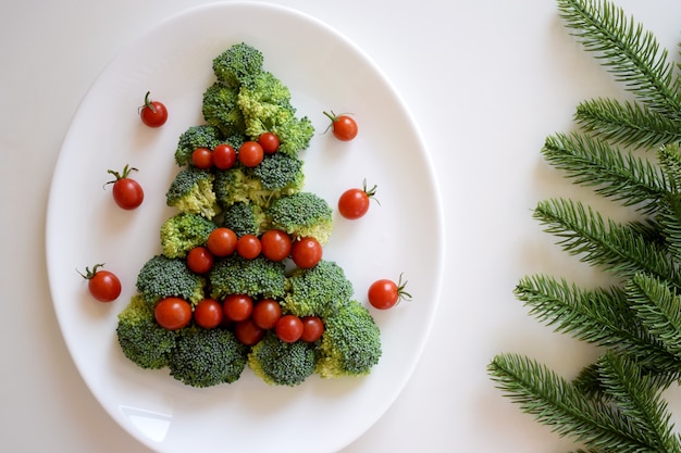 Christmas Tree made of broccoli and cherry tomatoes on white plate with fir tree branches on white background. Healthy organic food.