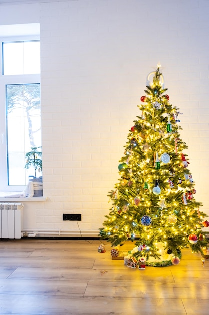 Christmas tree in the white interior of a house with large windows Glowing fairy lights garlands interior decoration of the studio room Potted plants in the home