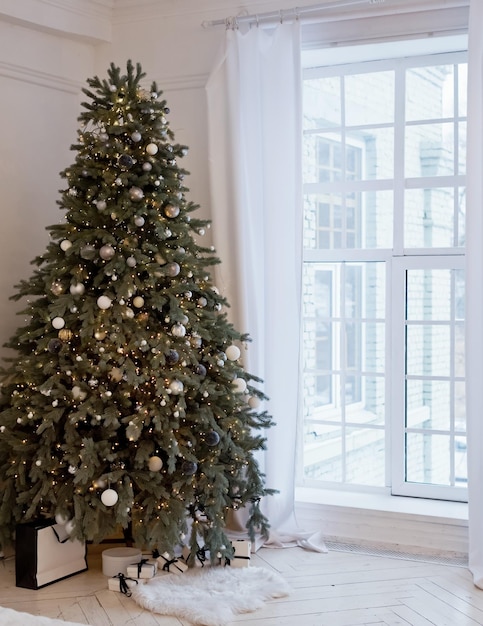 Christmas tree with decorations lights and gift boxes near big white window