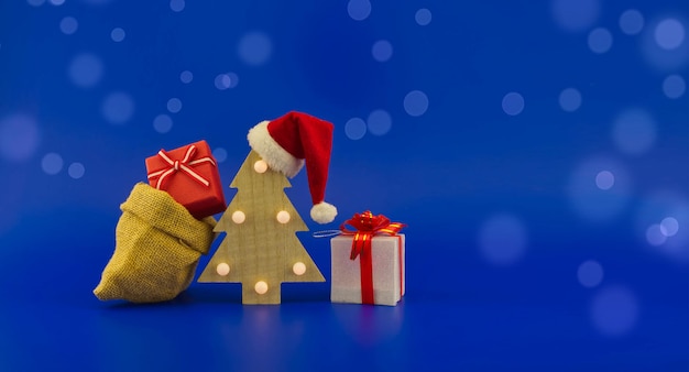 Christmas tree with red santa hat and jute sack with gift boxes\
on a festive blue background