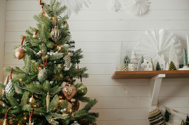 Christmas tree with vintage baubles and little christmas village decoration Modern christmas tree and stylish little trees and houses scene in festive decorated room Atmospheric Winter holidays